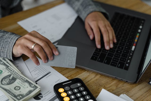 person-using-a-laptop-while-holding-a-bank-card-with-cash-documents-and-a-calculator-on-the-table person-using-a-laptop-while-holding-a-bank-card-with-cash-documents-and-a-calculator-on-the-table