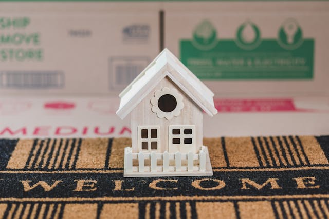 a-miniature-wooden-house-placed-on-a-welcome-doormat-with-moving-boxes-in-the-background a-miniature-wooden-house-placed-on-a-welcome-doormat-with-moving-boxes-in-the-background