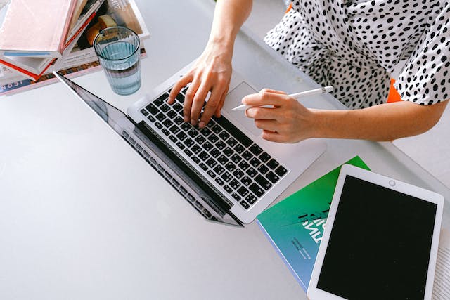 a-person-typing-on-a-laptop-with-a-stylus-in-hand-surrounded-by-books-a-glass-of-water-and-a-tablet-on-a-desk a-person-typing-on-a-laptop-with-a-stylus-in-hand-surrounded-by-books-a-glass-of-water-and-a-tablet-on-a-desk